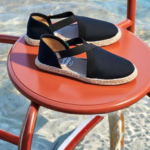 "Sandalia Flex Relic negra de tela con tiras cruzadas y suela de yute trenzado sobre fondo de agua."