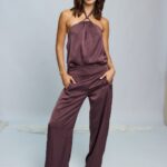 Modelo posando con la Blusa Galileo color ciruela de tela satinada. La blusa tiene cuello tipo halter con un herraje circular metálico en el centro del escote y caída fluida. Se combina con un pantalón de la misma tela y color."