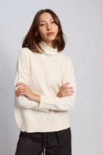 "Mujer vistiendo la Polera Anthony tejida en color negro, off white y gris melange, con cuello alto, hombros caídos y calce holgado sobre pantalón oscuro, marca Agustina Dominguez."