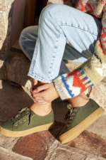 Persona sentada en escalones de piedra vistiendo zapatillas de lona verdes con cordones a juego y suela ancha de goma color marrón claro. Look casual con jeans y jersey de lana estampado."