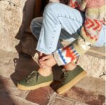 Persona sentada en escalones de piedra vistiendo zapatillas de lona verdes con cordones a juego y suela ancha de goma color marrón claro. Look casual con jeans y jersey de lana estampado."