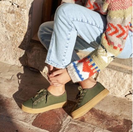 Persona sentada en escalones de piedra vistiendo zapatillas de lona verdes con cordones a juego y suela ancha de goma color marrón claro. Look casual con jeans y jersey de lana estampado."