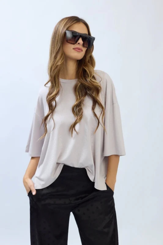 Fotografía de estudio de una modelo mujer posando con la Remera Duna blanca, de corte oversize, mangas cortas y hombros caídos. Viste pantalones cortos gris plateado y posa con las manos en los bolsillos sobre un fondo blanco neutro.